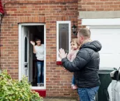 Father is carrying his baby daughter to the car and is turning round to wave at the mother who is stood at the front door of the house.