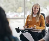A woman looks happy and confident as she leads a group discussion at her place of work. She is a wheelchair user and has Muscular Dystrophy.