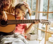 Adult woman at home during quarantine with her young daughter who is playing a guitar