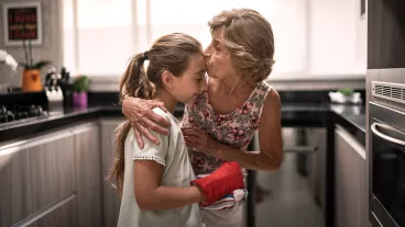 An older woman in the kitchen leans down to kiss a young girl on the forehead.